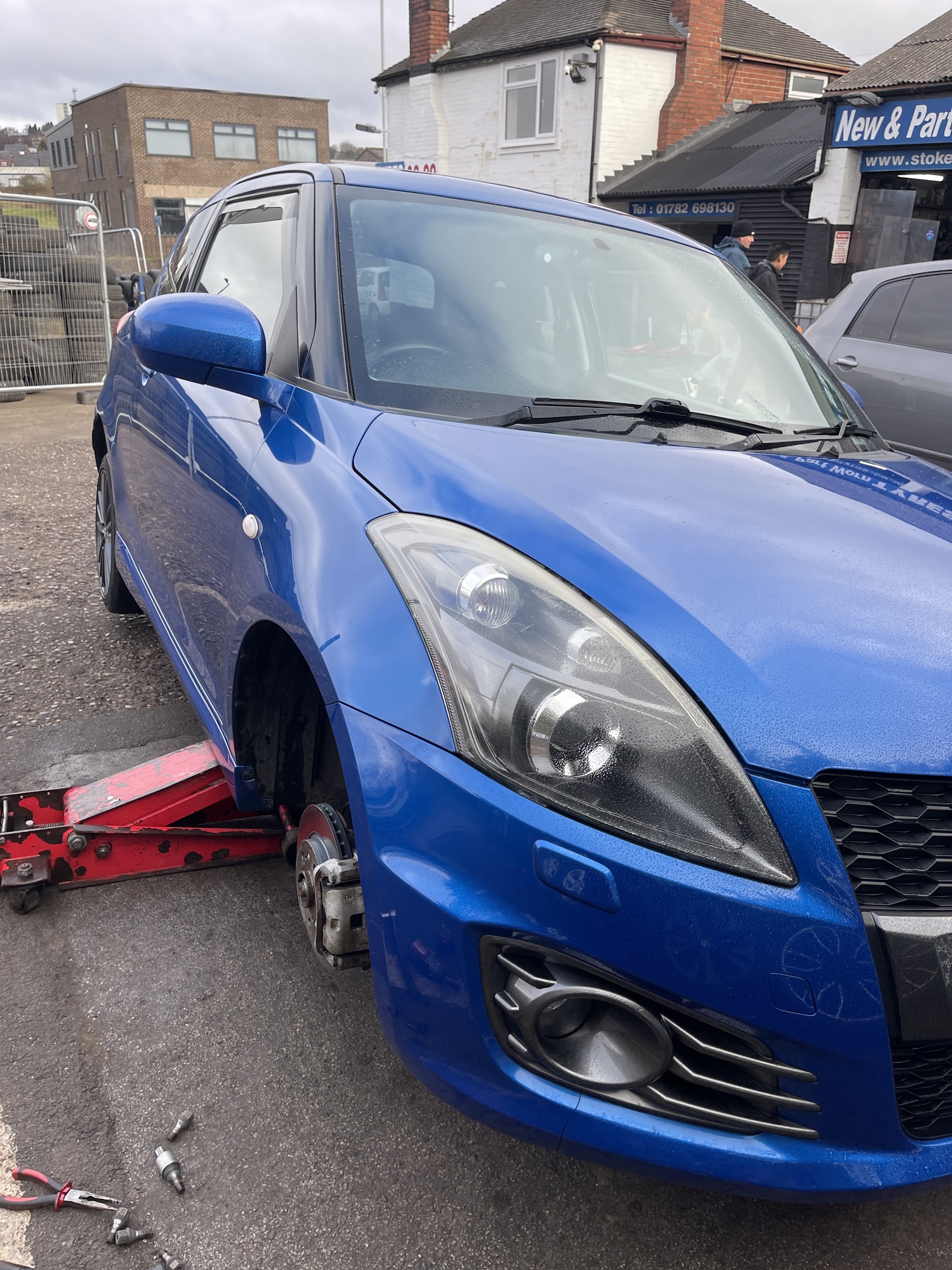 A blue Suzuki Swift on a trolley jack with its front wheel off on the Stoke Tyres forecourt