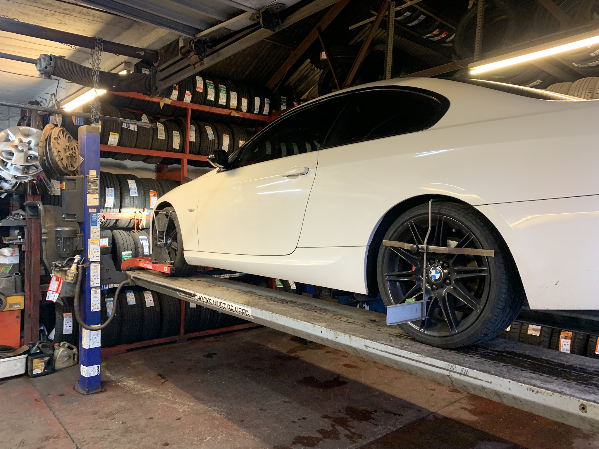 A white BMW up on the alignment lift in the Stoke Tyres workshop, with tracking clamps on its wheels and racks of new tyres behind